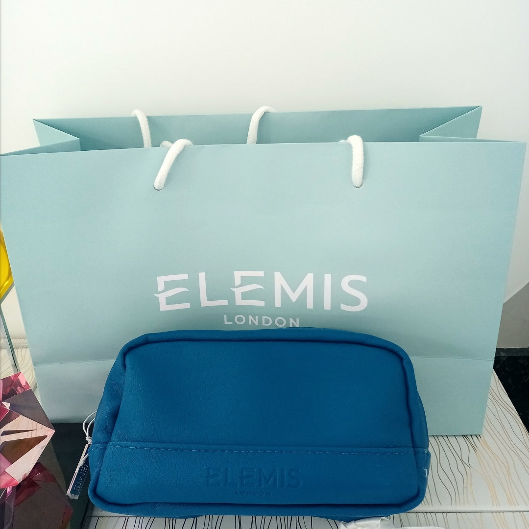 Blue Elemis pouch in front of a matching shopping bag on a light surface.