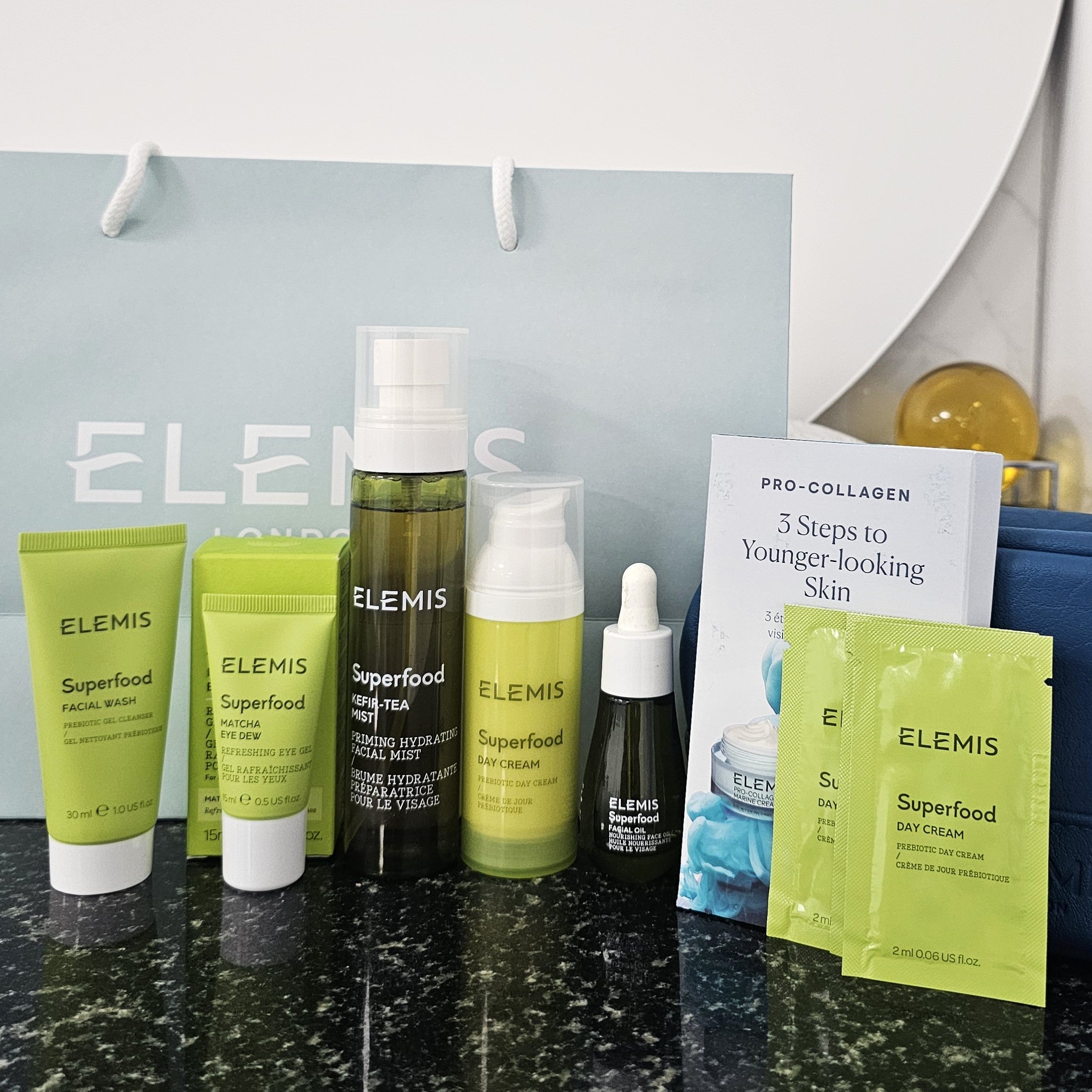 Elemis skincare products including bottles, tubes, and packets on a countertop with a branded bag in the background.